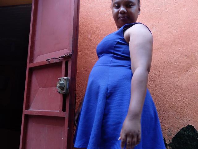 Leylahwilliam-hot, curvy black woman in a blue dress, standing near a red door, looking down with a slight smile, showing her ass, and one arm extended forward.