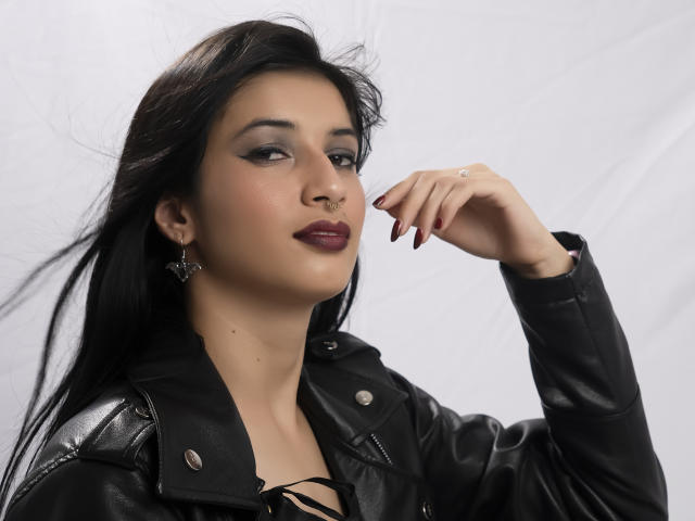 AlisKooper with dark hair, black leather jacket, nose ring, smoky eye makeup, maroon lipstick, ring on finger, and black earrings, posing with hand near face.