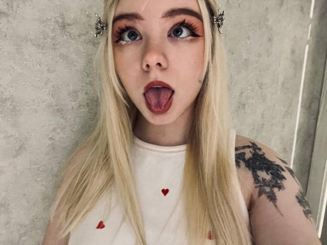 EnchantingEyes with blonde hair, blue eyes, and a tattoo on her right arm, tongue out, wearing a white shirt with red hearts, and butterfly hair clips.