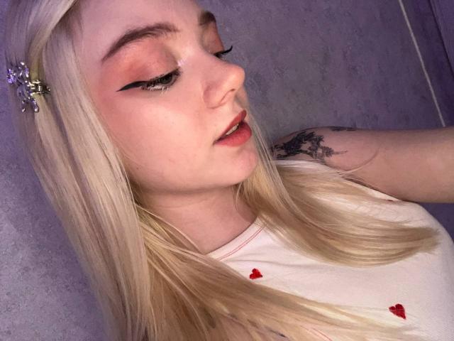 EnchantingEyes has blonde hair, pink eyeshadow, black eyeliner, and a small nose. She's wearing a white shirt with red hearts, and has a tattoo on her right arm.