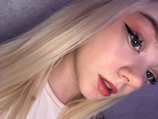 EnchantingEyes has blonde hair, pink eyeshadow, black winged eyeliner, red lips, and blue eyes. She's looking at the camera with a slight, sexy, open-mouthed smile.