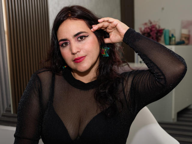 KathyFlores with dark, wavy hair, wearing a sheer black top with sparkly fabric, showing her big tits in a black bra, pink eyeshadow, and red lipstick.