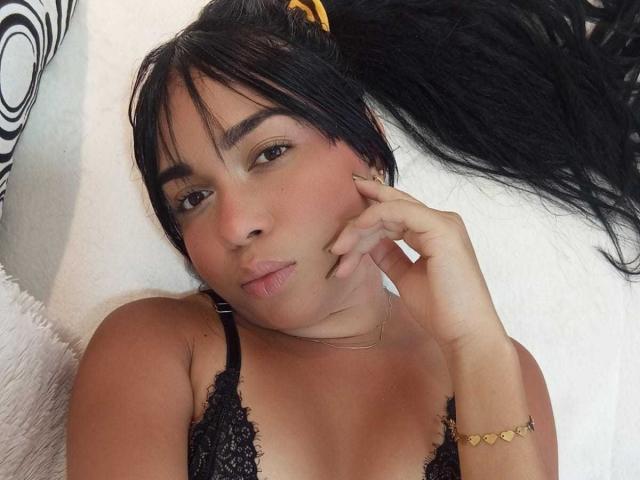 Alice Smity, con un reggiseno nero in pizzo, labbra piene, occhi marroni, lunghi capelli neri raccolti in una coda di cavallo, un braccialetto dorato e una collana, sdraiata su un cuscino bianco, che tocca la guancia con la mano..