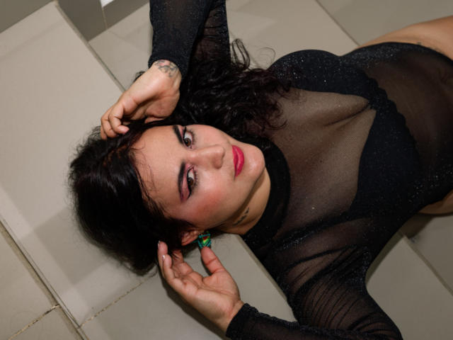 KathyFlores with dark hair, red lipstick, and black sheer dress, lying on tiled floor, arms above head, looking at camera, visible tattoo on left wrist, turquoise earrings.
