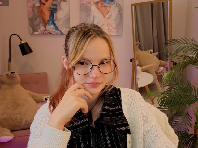GingerEmma avec des lunettes, des cheveux blond-rosés, un cardigan blanc, une robe noire à rayures fines, une main sur le menton, un sourire discret, un mur rose, un miroir, une lampe, un ours en peluche, une plante en pot, des tableaux..
