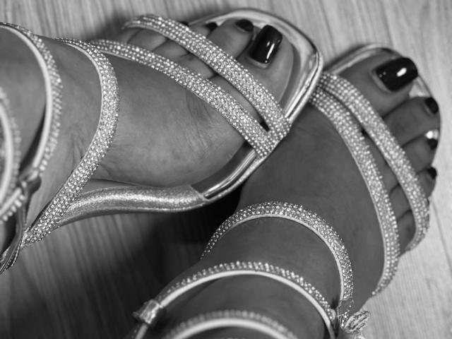 LillyTaylor's feet in sparkly silver high heels with glittery straps, showing dark purple toenail polish. Her toes are slightly curled. Wood floor underneath.