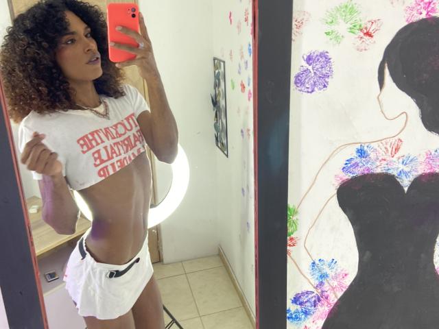 Jhoanesil with curly brown hair, wearing a white crop top and shorts, showing a slim, toned, dark-skinned body, holding a red phone in a mirror selfie.