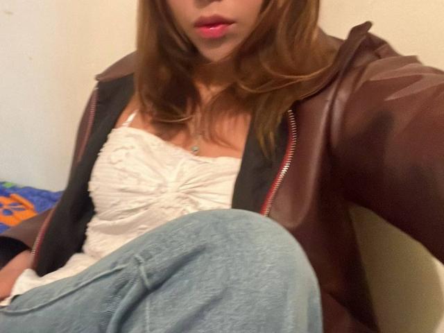 HeraJuno con i capelli castani, le labbra rosa, una giacca di pelle marrone, una camicetta bianca con volant e jeans blu, seduta con le gambe incrociate, mostrando un piccolo sprazzo del seno..