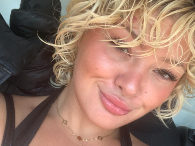 Le visage de DivineBlondee, avec ses cheveux blonds bouclés, sa peau légèrement rosée, ses lèvres roses et son sourire discret. Elle porte un haut noir et un collier en or..