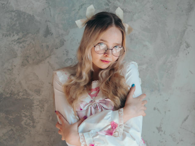 BellaMoor with wavy blonde hair, wearing glasses, a white dress with pink bows, and white ribbons in her hair, hugging herself with blue and pink nails.