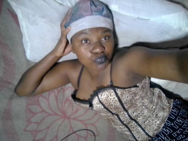 A photo of HabbyBaboy lying on a bed. HabbyBaboy has dark brown skin and is wearing a patterned headscarf and a leopard-print corset with black straps. HabbyBaboy's right hand is on the headscarf, and they have a neutral expression with slightly parted lips. The corset's texture is shiny and detailed. The background includes a white pillow and a beige carpet with a pink floral pattern. The image has a casual, slightly disordered feel.