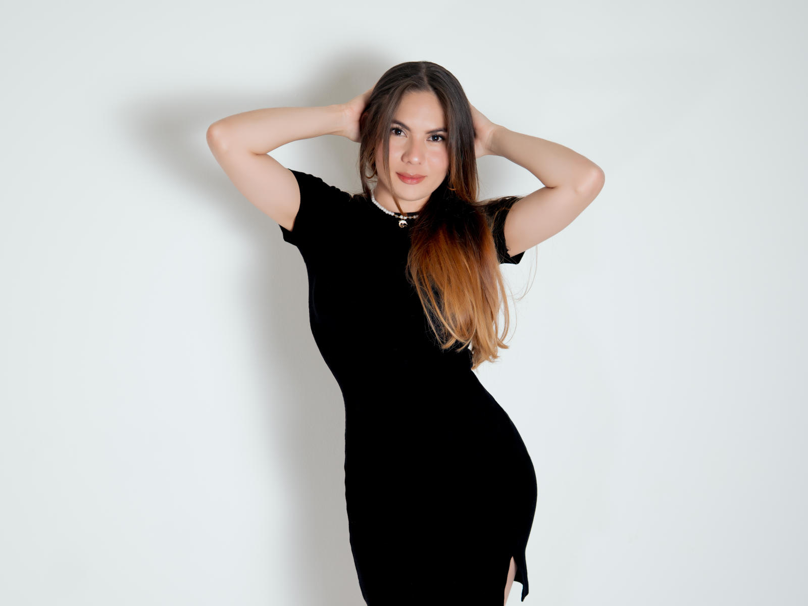MaddySimons steht mit den Händen hinter dem Kopf, trägt ein eng anliegendes schwarzes Kleid, langes braunes Haar und eine schwarze Halskette. Dabei zeigt sie ihren Hintern und hat ein leichtes Lächeln..