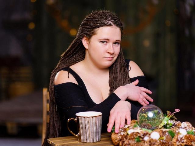 JanesBondi with long braids, black off-shoulder top, looking at camera, hands on table, mug, glass dome, and wreath in front, subtle smile, brown eyes, cute, medium tits.