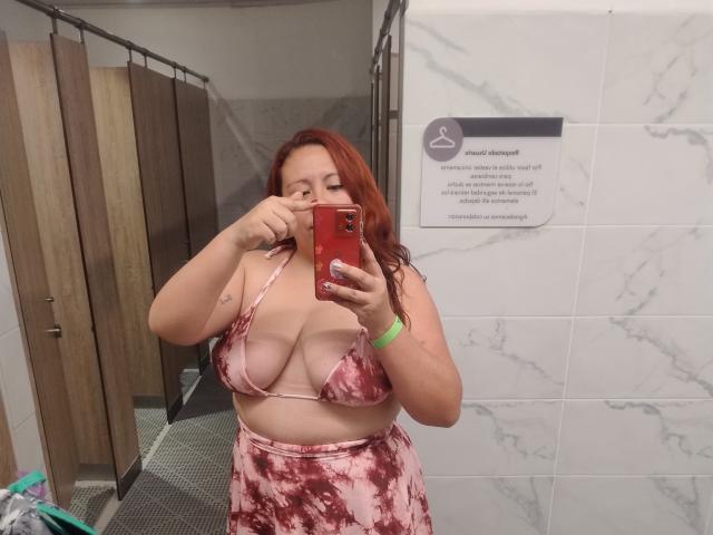 LexyDom with red hair, wearing a pink tie-dye bikini top, showing her tits, and matching skirt, taking a mirror selfie in a bathroom, biting her finger.