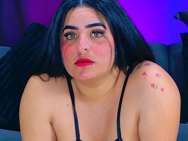KatrinaStones has black hair, red lipstick, and a tattoo of small red hearts on her right shoulder. She's wearing a black strap dress, looking at the camera.