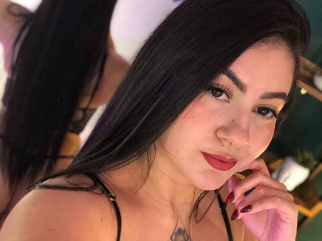 AlanaaWalker with black hair, red lipstick, and a nose ring, looking at the camera, showing her tits with a black strappy top, red nails, and a tattoo on her chest.