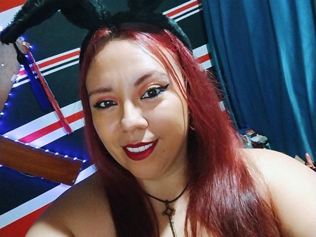 LexyDom with red hair, black bunny ears, and red lipstick, smiling at the camera, showing her tits, wearing a black necklace with a cross.