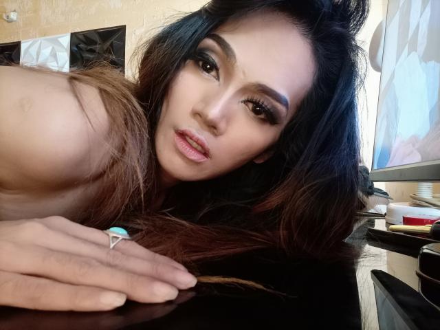 HugeCockJackie, with long brown hair and makeup, leans on a black surface, showing off her pouty lips and ring on her hand. Her tits are partially visible.