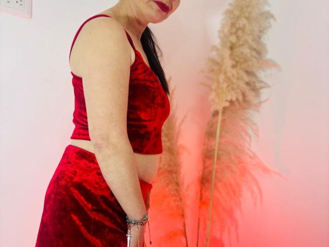 DolcceFerrer trägt ein rotes Sammt-Crop-Top und einen passenden Rock, hat blasse Haut, schwarzes Haar, roten Lippenstift, hält flauschige, beige Federn, ein rotes Armband am Handgelenk, ist im Profil zu sehen und lächelt..