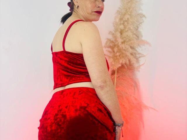 DolcceFerrer in rode fluwelen lingerie, met de rug naar de camera, waarbij haar achterkant zichtbaar is, met een pluizig veertje en rood licht. Ze heeft donker haar, rode lippenstift en een zilveren armband..