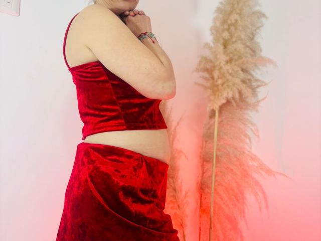 DolcceFerrer with pale skin wearing a red velvet crop top and matching skirt, hands near mouth, standing next to a puffy beige feather. Red light on right.