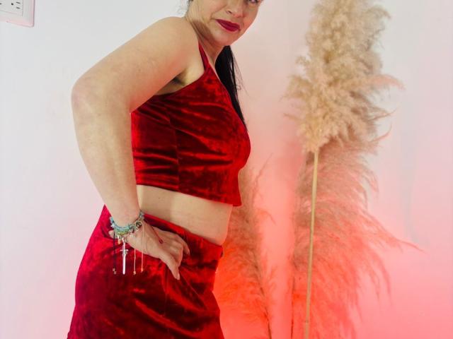 DolcceFerrer in red velvet crop top and matching skirt, red lipstick, hand on hip, holding keys, with puffy beige grass in background, smiling slightly.