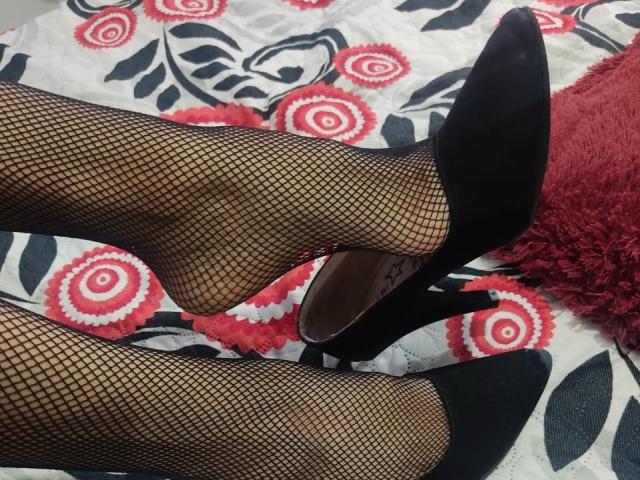 Danny69s' Beine in schwarzen Fischstrumpfhosen und schwarzen High Heels mit spitzer Spitze, die eine offene Ferse zeigen, auf einem gemusterten Bett. Ein rotes, flauschiges Kissen auf der rechten Seite..
