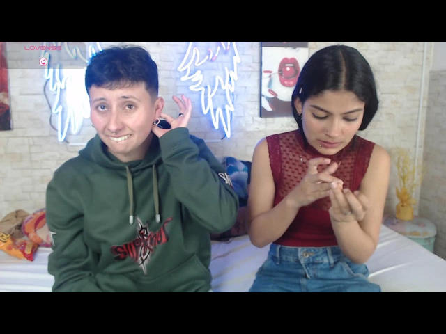 GabiAndMei with short brown hair in a green hoodie and another GabiAndMei with black hair in a red sleeveless top, focused on her hands. Bedroom, bed in background.