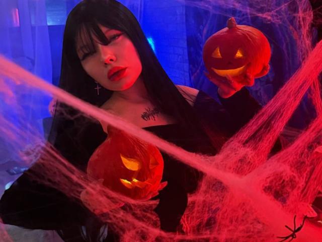 AmeliaAley with black hair, red lipstick, and a spider tattoo, holding two glowing jack-o'-lanterns, surrounded by red spiderwebs in a blue-lit room.