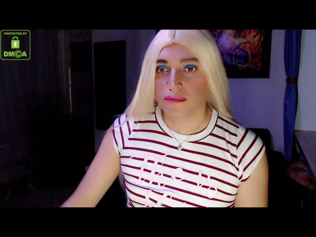 SamaraBlue with platinum blonde hair, blue eye makeup, pink lipstick, and striped white-red shirt, sitting in dim light, showing her tits slightly through shirt. Watermark in top left.
