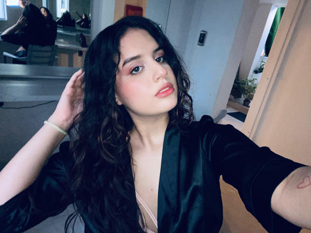 KylieLumi, con cabello negro largo y ondulado, vestida con una bata de satén negro, lencería rosa y lápiz labial rosa, sosteniendo su cabello, mirando a la cámara, con una ligera sonrisa..