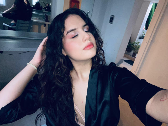 KylieLumi with wavy black hair, pink eyeshadow, and red lipstick, wearing a black satin robe, hand in hair, eyes closed, pink bra visible, white bracelet on wrist.