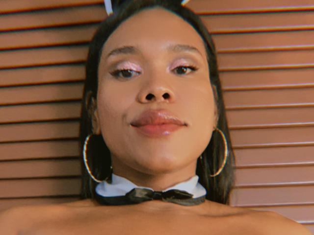 AshleyRob has brown skin, pink glittery eyeshadow, and glossy lips. She's wearing a black bowtie and large gold hoop earrings. Her nose is centered, and she's smiling slightly.