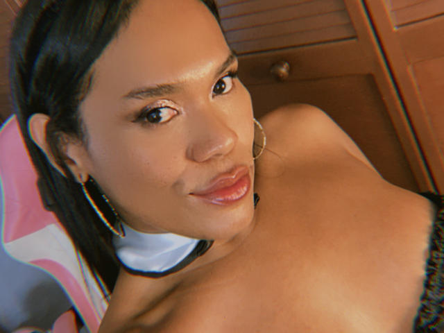 AshleyRob with shiny brown skin, black hair in a ponytail, hoop earrings, and glossy pink lips, topless, showing her small, round tits, smiling at the camera.