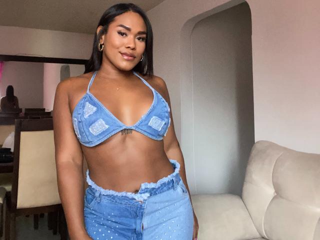 AshleyRob with brown skin, long black hair, wearing a blue denim bikini top and matching high-waisted jeans, showing her small tits and cute belly button tattoo.