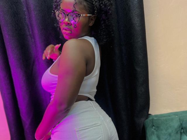 SexyBerlin with curly hair, wearing heart-shaped glasses, white tank top, and white jeans, showing her big ass, standing with pink and purple lighting on her back.