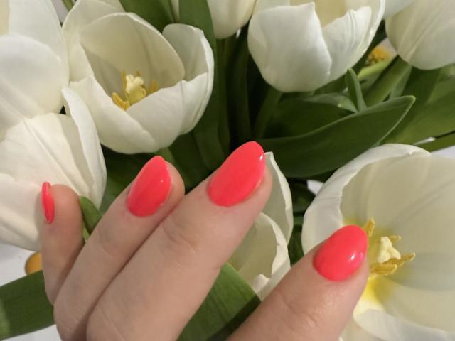 Elionoria's hand with bright pink, glossy nail polish holding white tulips, showing off her shiny, pointed nails against the soft petals and green leaves.