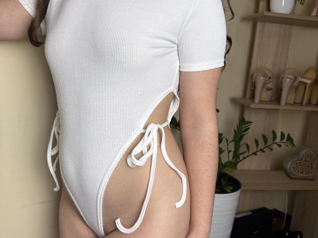 Symphoniia in a white ribbed one-piece swimsuit with side ties, showing her small tits and bare ass. Her arm is by her side, her skin is fair.