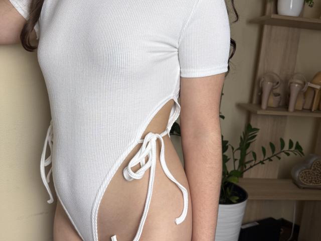 Symphoniia's white ribbed one-piece swimsuit with side ties, showing her bare ass and a side of her small, perky tits. Her arm is visible, and she's standing.