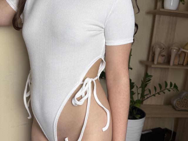 Symphoniia's white ribbed one-piece swimsuit with side ties, showing her bare ass and upper thighs. Her arm is visible, and she has light skin and brown hair.