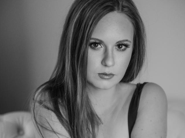 Black-and-white photo of LyraFantasy with long, straight, dark hair, staring intensely, wearing a black tank top, showing her tits, and a serious expression, lips slightly parted.