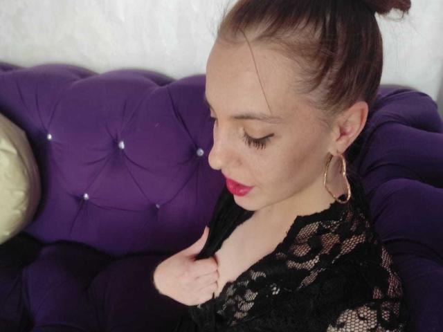 SexiMillie with brown hair in a bun, wearing a black lace top, gold hoop earrings, and red lipstick, sitting on a purple tufted couch, gently touching her chest.
