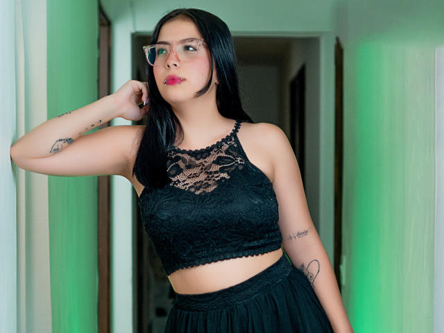 CherryLusts con top corto in pizzo nero, che mostra il seno, gonna nera, occhiali, tatuaggi e un corridoio illuminato di verde, con una mano sulla testa, che guarda la fotocamera..