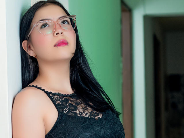 CherryLusts met een zwarte kanten top, roze bril en roze lippen, leunt tegen een muur. Ze heeft lang zwart haar en een schattig, sexy gezicht met subtiele make-up..