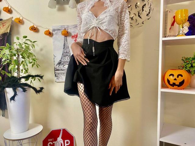 Millinia con una parte superior de encaje blanco, falda negra, medias de rejilla y tacones negros, mostrando su trasero y su vulva a través de la falda. Calabazas y plantas en el fondo..