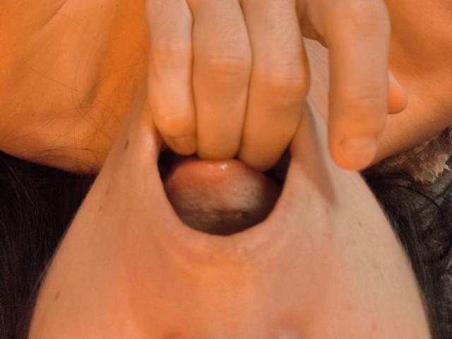 OrianaLaFrancaise's mouth is being held open by two fingers, showing her pink tongue and slightly visible teeth. Her face is partially visible with dark hair.
