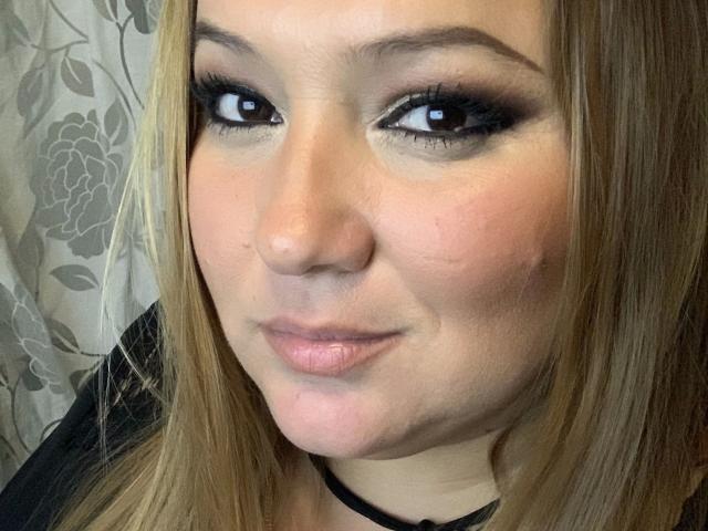 LaVa24-hot a un visage mignon avec des cheveux bruns clairs, un maquillage des yeux fumé et des lèvres roses. Elle porte un collier noir et a un léger sourire. Ses seins sont petits..