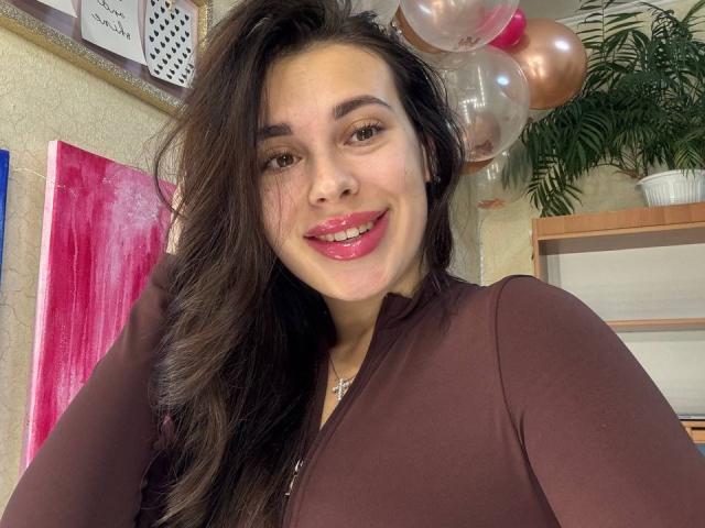 BlackFatal met lang donker haar, bruine ogen en roze lippenstift, gekleed in een bruin shirt met lange mouwen, lachend naar de camera met ballonnen op de achtergrond..