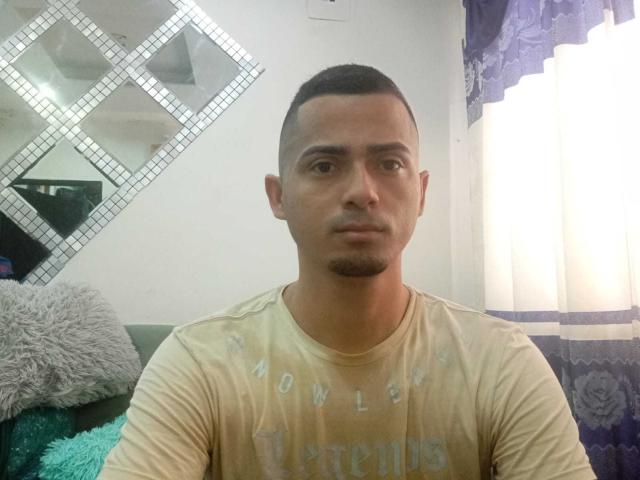 SamuAndHarrison tiene un tono de piel medio, cabello negro corto y un bigote. Lleva una camiseta amarilla y está sentado con una expresión neutra. Sus glúteos no son visibles..