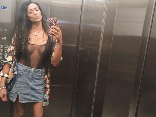 DiannaBanana with long black hair, wearing a sheer black top revealing small tits, a colorful floral kimono, and a blue denim skirt with a front slit, taking a selfie.
